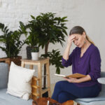 Upset beautiful 60 year old woman sitting on gray chair in living room, touching forehead and looking at open copybook on her lap, feeling frustrated because she forgot about important meeting