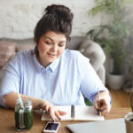 Hardworking positive young female freelancer with heavy curvy body multitasking at home office, texting sms on blank screen mobile phone and making notes in her diary, writing down plans and ideas